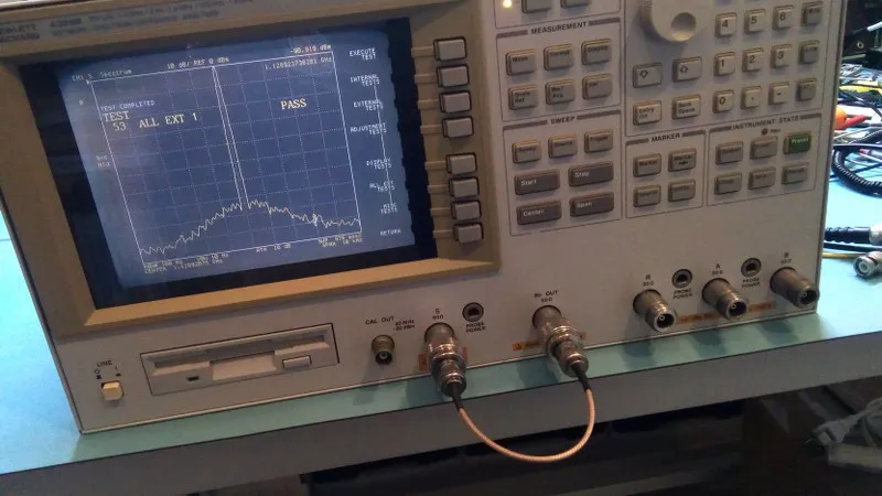 A coaxial cable, likely RG316 or RG174, connected to a vector network analyzer (VNA) or signal testing equipment on a lab bench. A coaxial cable, likely RG316 or RG174, connected to a vector network analyzer (VNA) or signal testing equipment on a lab bench.